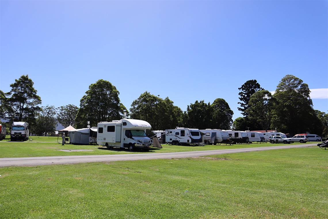 A picture of the campground with a number of camper vans, tents and trailers parked on the grass sites. There are trees in the background, and a green lawn in the foreground.