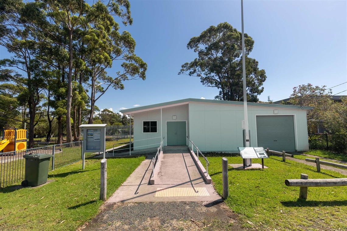 A view of the progress hall from the street. There is a green front lawn and a pathway leading to the blue community hall building, and a playground off to the left.