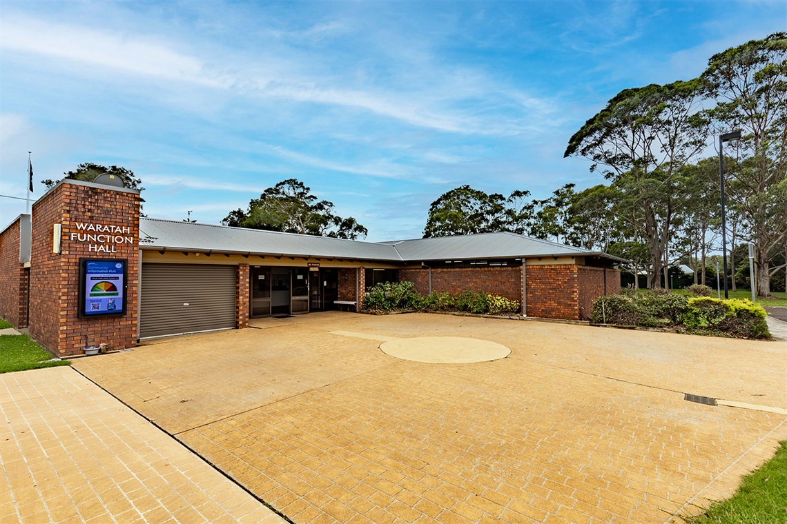 The exterior of the Waratah Function Hall. There is a large paved driveway in front of the building, and a community information hub under the building sign. There is a large entryway with glass double-doors.