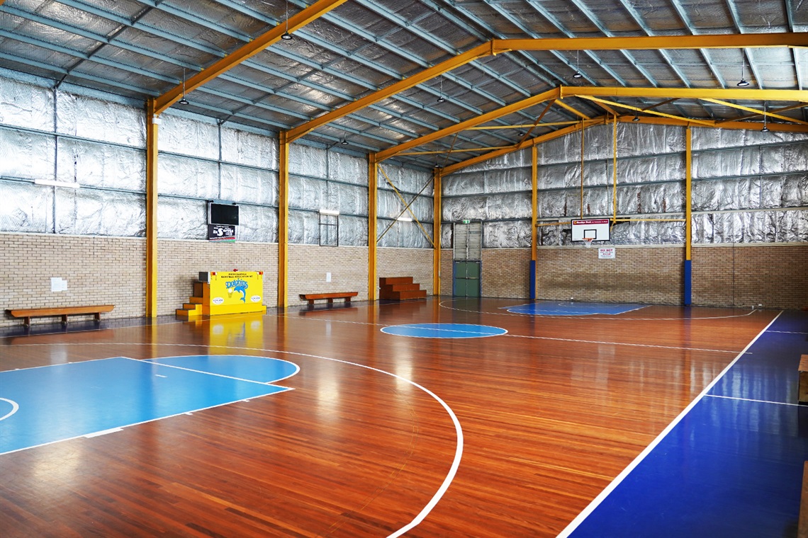 The Southern basketball stadium court. It has polished wood floors and a steel roof with reflective insulation along the walls. The bottom half of the walls is made of brick and there are benches up against the wall. 