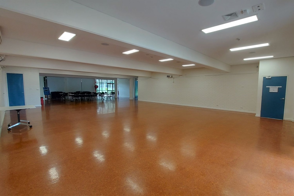 Indoor auditorium on lower ground level, tan wood flooring, doors at both ends onto grounds.