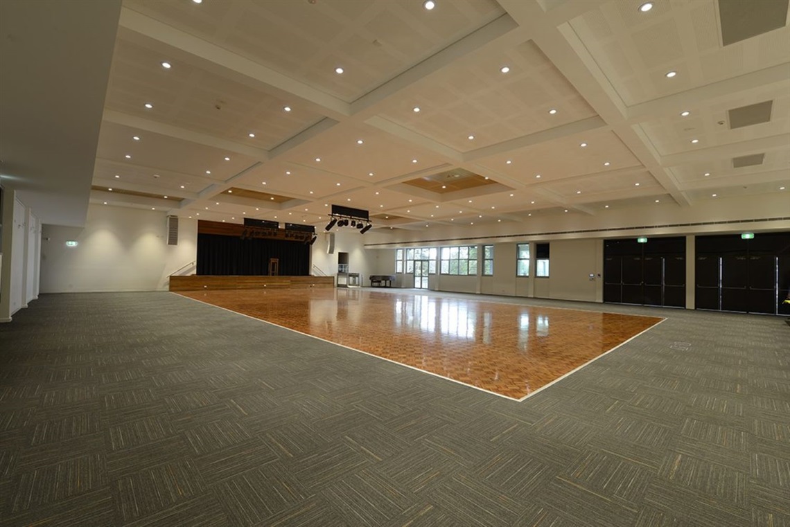 Large auditorium, parquetry flooring surrounded by grey carpet, stage with lighting and AV, dimmable ceiling lights, door to balcony at far end.