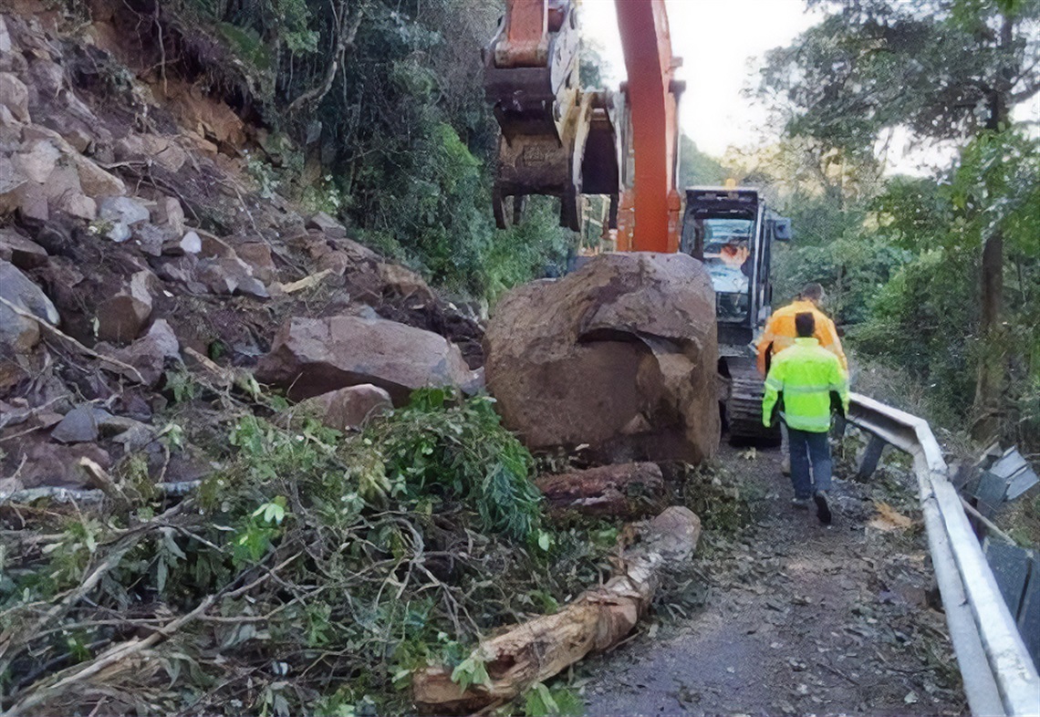 A photo of a large boulder and broken trees blocking a road, with an excavator behind the boulder and workers walking around the side of it.