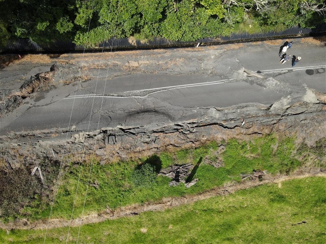An aerial view of a large section of road that has slipped down a hill, causing a large rift in the road.