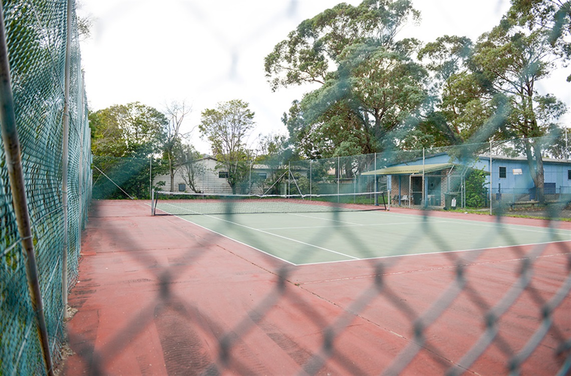 Single concrete tennis court surrounded by fencing and bordered by trees in the background