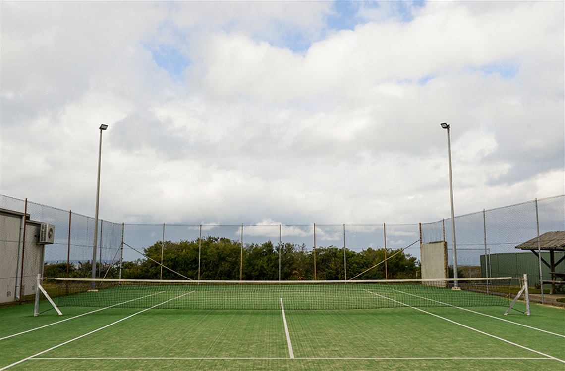 Single synthetic tennis court surrounded by fencing and bordered by Community Hall and sheltered area