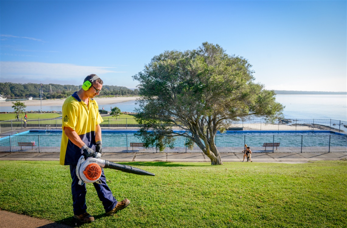 Shoalhaven City Council employee in high vis using a leaf blower to clear a grassed area in front of Huskisson ocean pool