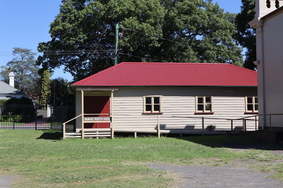 The front of the Rural Youth Hall building. There is a small awning in front of the entrance double doors. There is a ramp leading up to the doors on one side, and small set of stairs on the other. The Pavilion building can be seen to the right, and Albany Street in the background behind the fence.