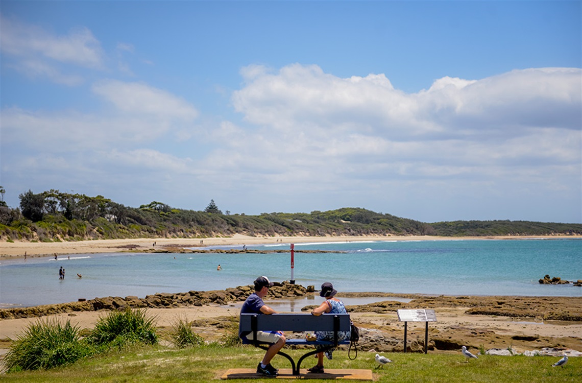 Two people sitting on bench seat overlooking a rivers entrance to the ocean with calm water