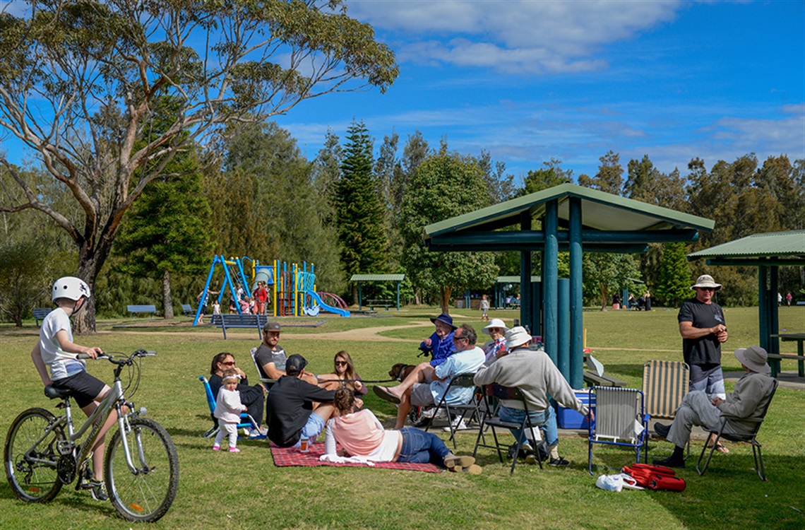 Family group picnicing in park alongside shaded bbq areas and play equipment