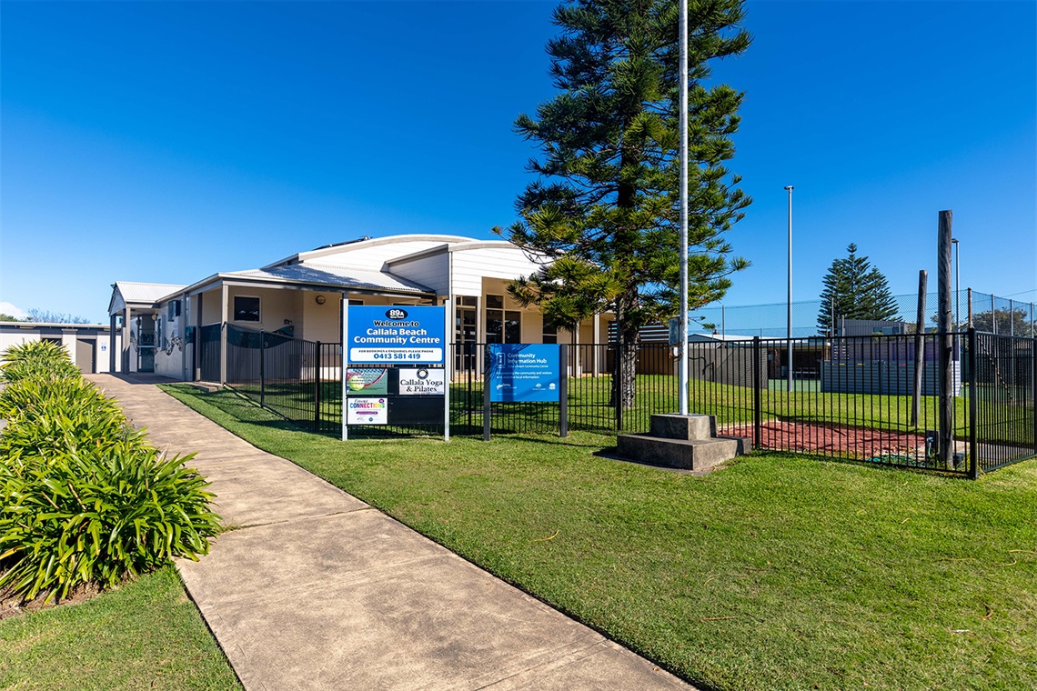 The view of the Callala Beach Community Centre from the footpath. There is a concrete walkway leading up to the building, and a fenced-in courtyard at the front. A tennis court can be seen in the background. 