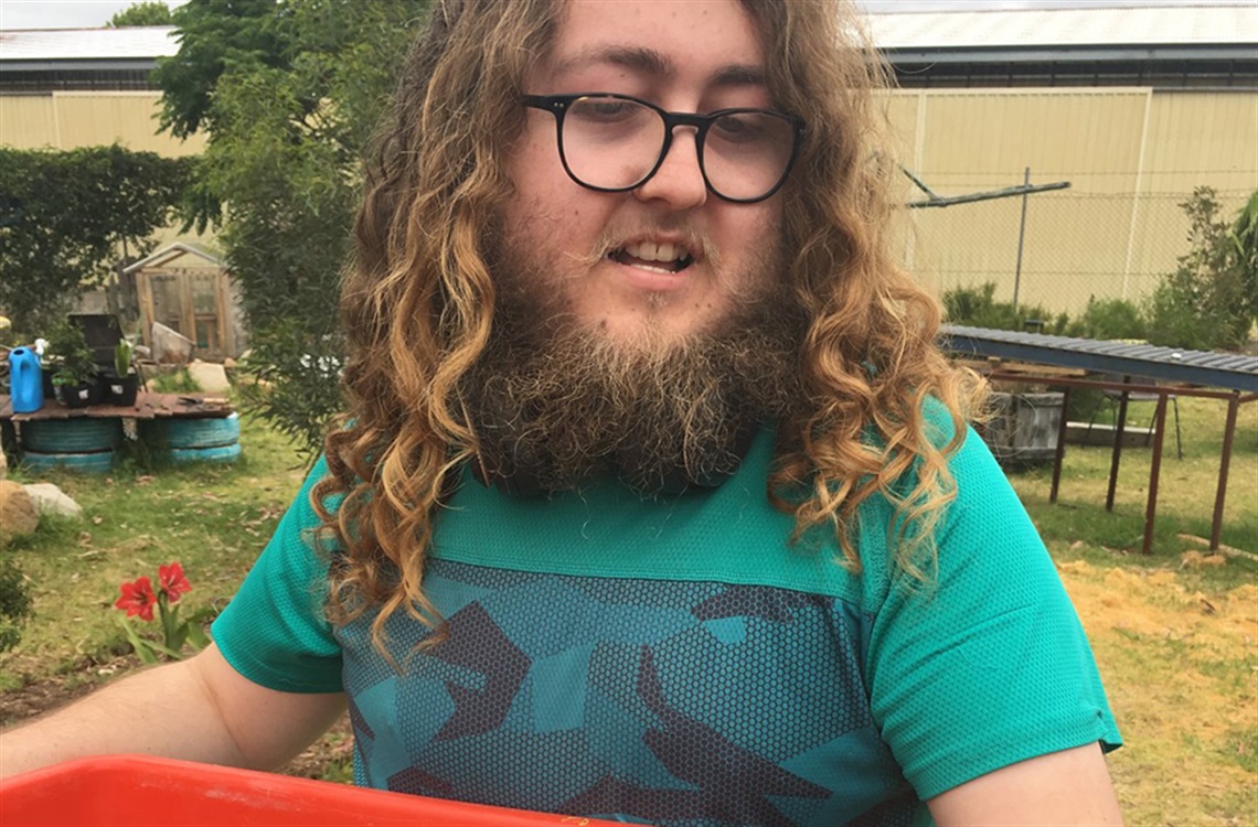 Young person with glasses and long hair holding orange tub in community garden